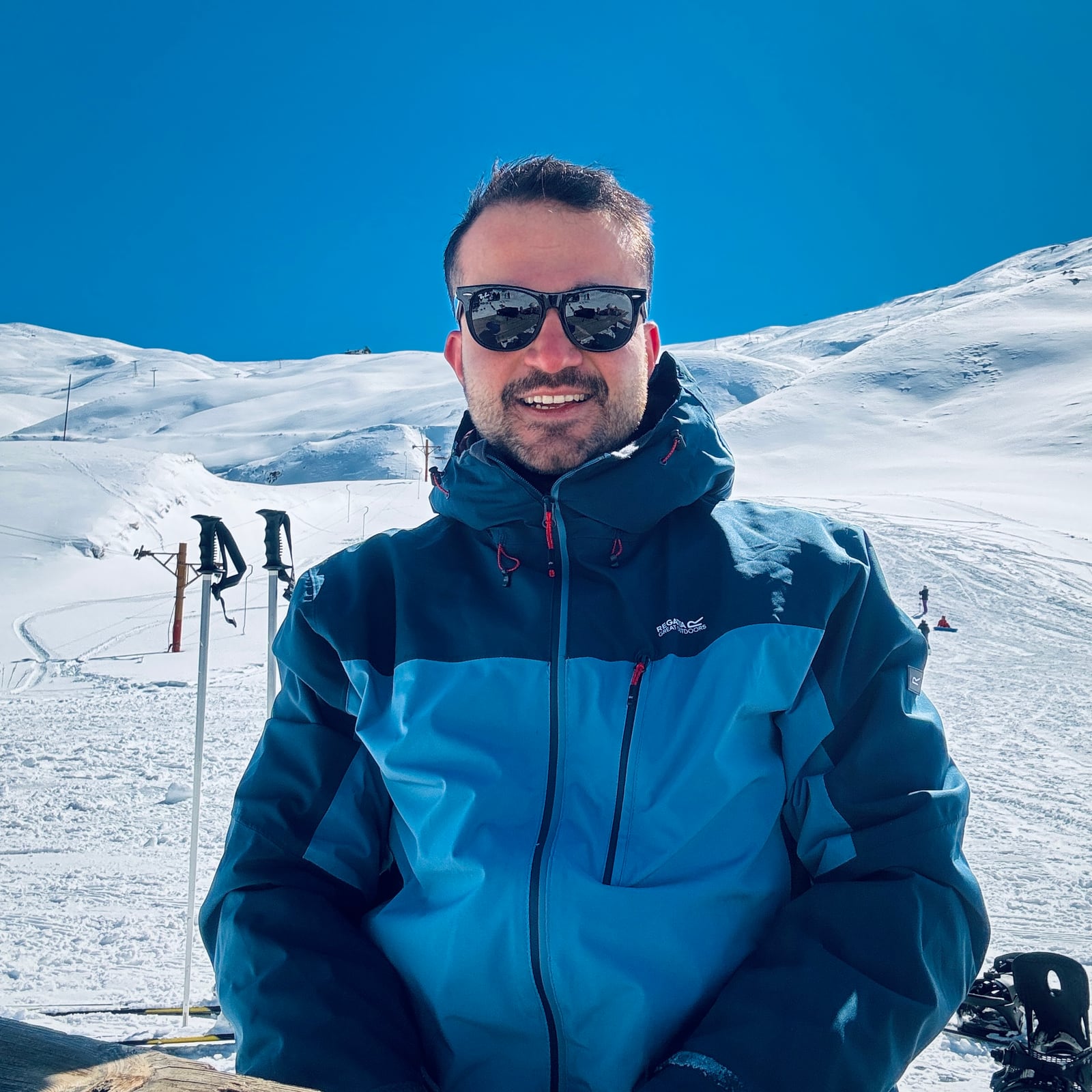 Omid Mirzaei (Omid Hybrid) smiling at a ski resort in a blue jacket and sunglasses